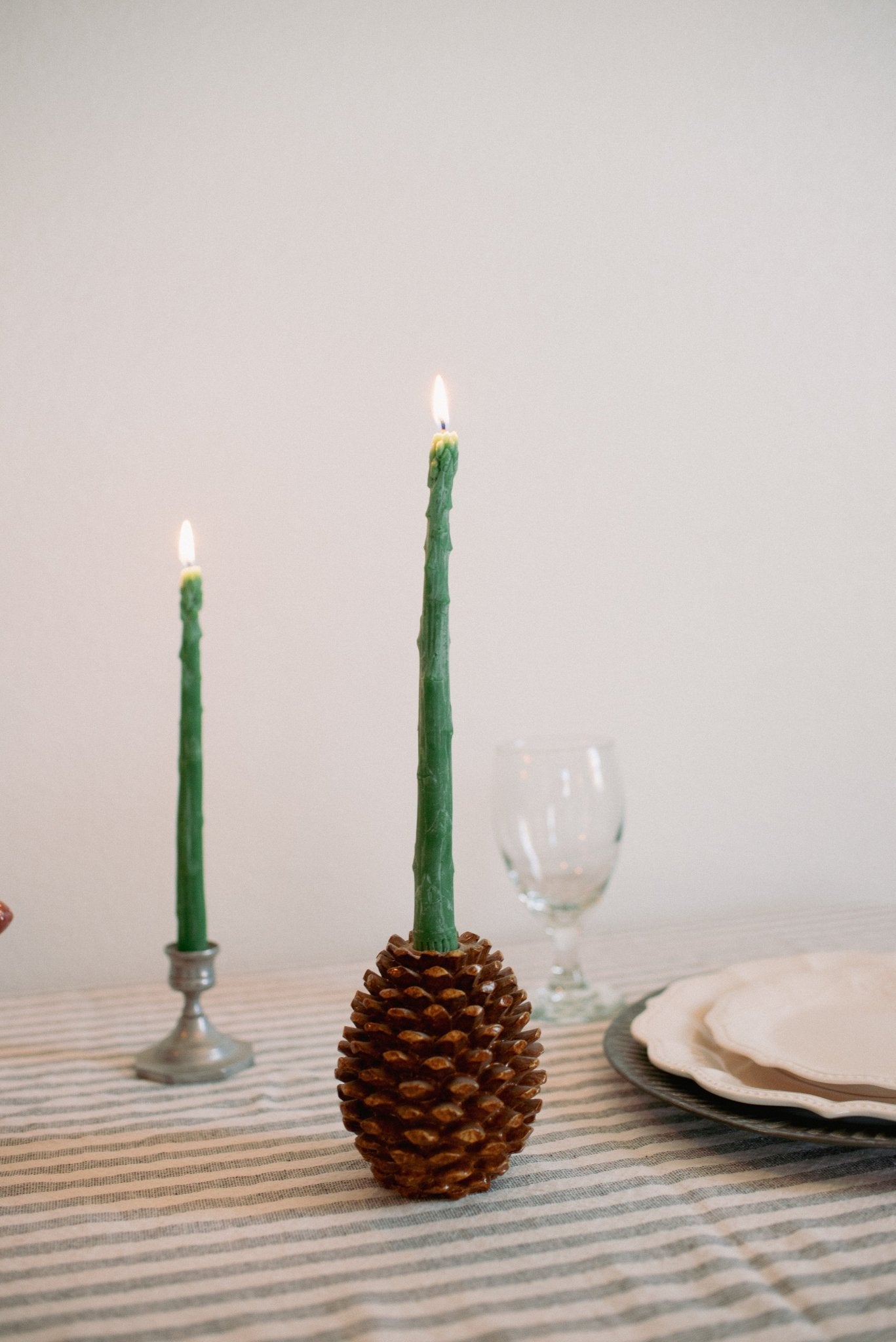 Green candle in a pine cone holder on a table with other candles and a glass.