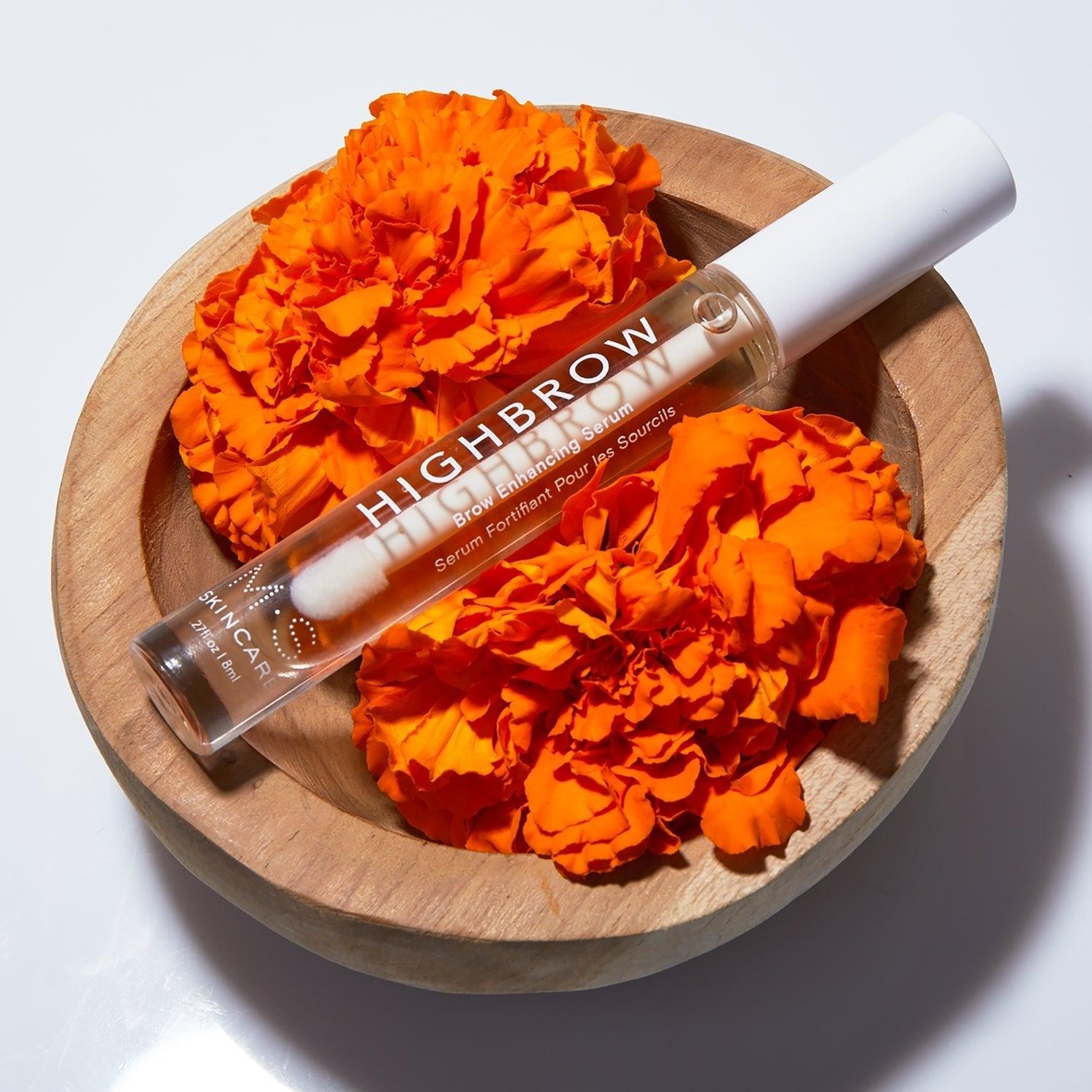 M.S Skincare HIGHBROW Brow Enhancing Serum in a glass container with white wand & cap on top of a wood bowl with marigolds.